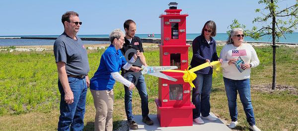 5 people stand next to a red Little Free Library that's shaped like a lighthouse with 3 windowed doors holding books. A yellow ribbon is being cut with a giant scissors in front of it.
