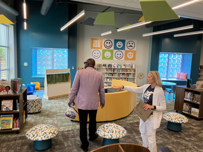 Dr. Williams is being shown around the children's area in the library by a staff member. The area is full of colorful chairs, book shelves, tables, and large colorful windows in the background.