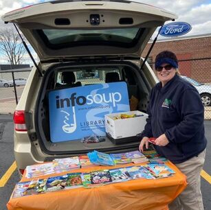 Smiling library staff standing next to a table full of books and oversized InfoSoup library card in an open van trunk
