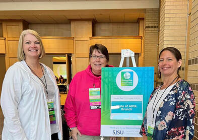 Paige, Cathy & Kristie smile standing in front of ARSL sign