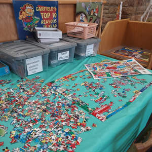 jigsaw table and containers of magnetic tiles, snap circuits, sketch pad and more on top of a covered table in the library