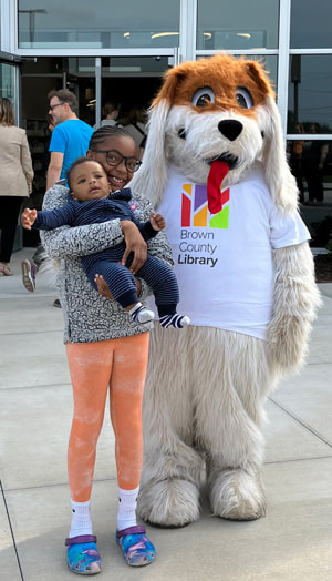 A child is holding an infant and smiling and standing next to Rover Reader (someone in a dog costume and wearing a Brown County Library T-shirt).