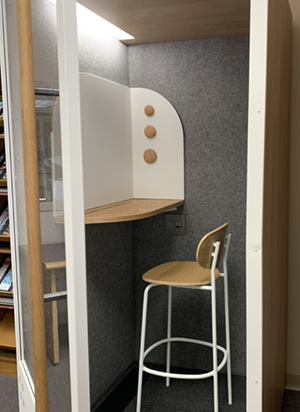 This is a picture of the booth with the door open, showing a lit interior with woodgrain ceiling, gray interior fabric walls, and a tall white and woodgrain chair a chair and matching workspace mounted on the wall with two white magnetic wall sections. There's also a plug-in spot on the wall below the workspace.