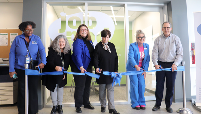 6 adults, all with scissors, cut a blue ribbon in front of a JobPod workspace with a glass front