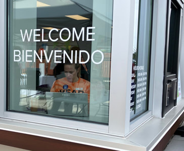 A library staff member is using a laptop, behind a window that says "Welcome/Bienvenido" 