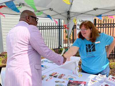Dr. Williams is shaking hands with a smiling library staff member at a table full of giveaways and information, all under a canopy outside.