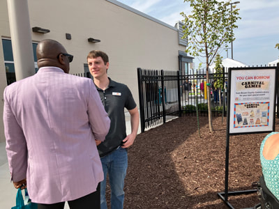 Dr. Williams talking to staff member Jerad. They're standing in front of a sign advertising how patrons can borrow carnival games from the library.